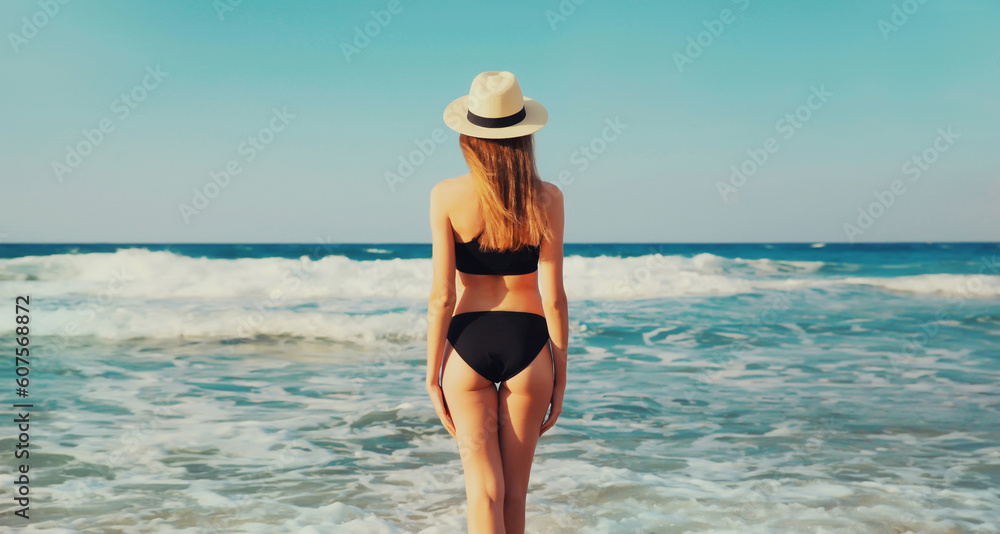 Summer vacation, rear view of beautiful young woman in bikini swimsuit and straw hat on the beach on sea coast with waves background