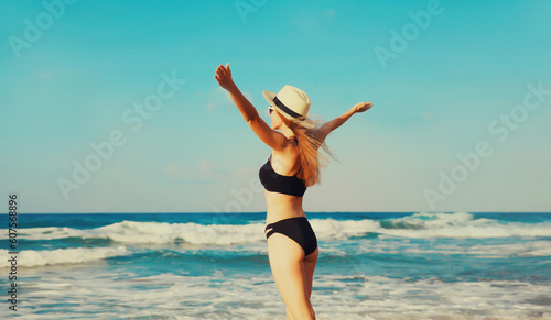 Summer vacation, rear view of beautiful happy slim woman in bikini swimsuit and straw hat raising her hands up on the beach on sunny day on sea coast background