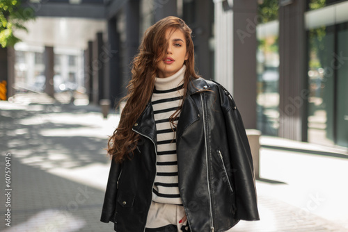Beautiful young fashionable brunette girl in trendy clothes with a stylish leather jacket and a striped sweater walks in the city on a sunny day