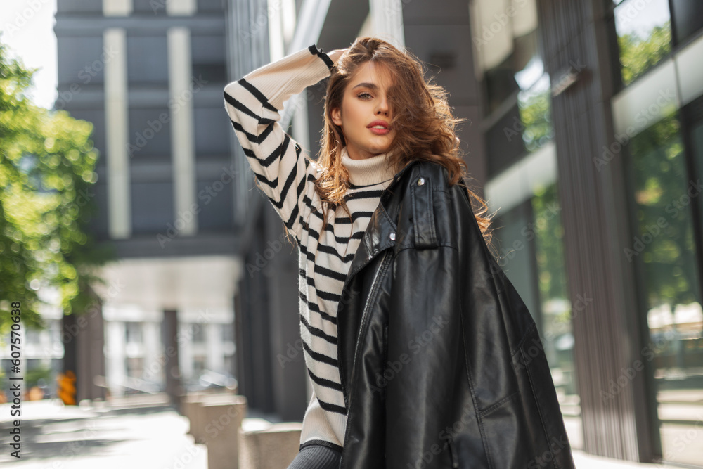 beautiful young fashion girl in stylish casual clothes with a leather jacket and a striped sweater walks and poses on a sunny day in a modern city