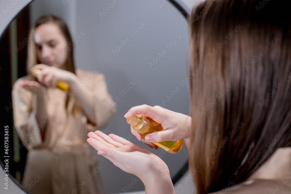Yong Woman Brunette Applying Essential Oil Spray On Her Hair Standing Near Mirror In Bathroom. Split Ends Repair Treatment. Concept of protection and care for hair Yong Woman Brunette Applying Essential Oil Spray On Her Hair Standing Near Mirror In Bathroom. Split Ends Repair Treatment. Concept of protection and care for hair