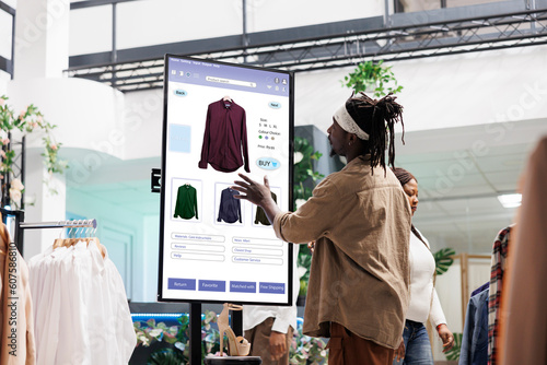 Customer using touch display shopping for fashion items in clothing store, interactive board. African american man choosing stylish clothes with different designs, modern kiosk service. photo