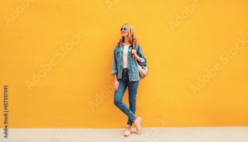 Full length beautiful smiling young woman wearing denim jacket, backpack on yellow background