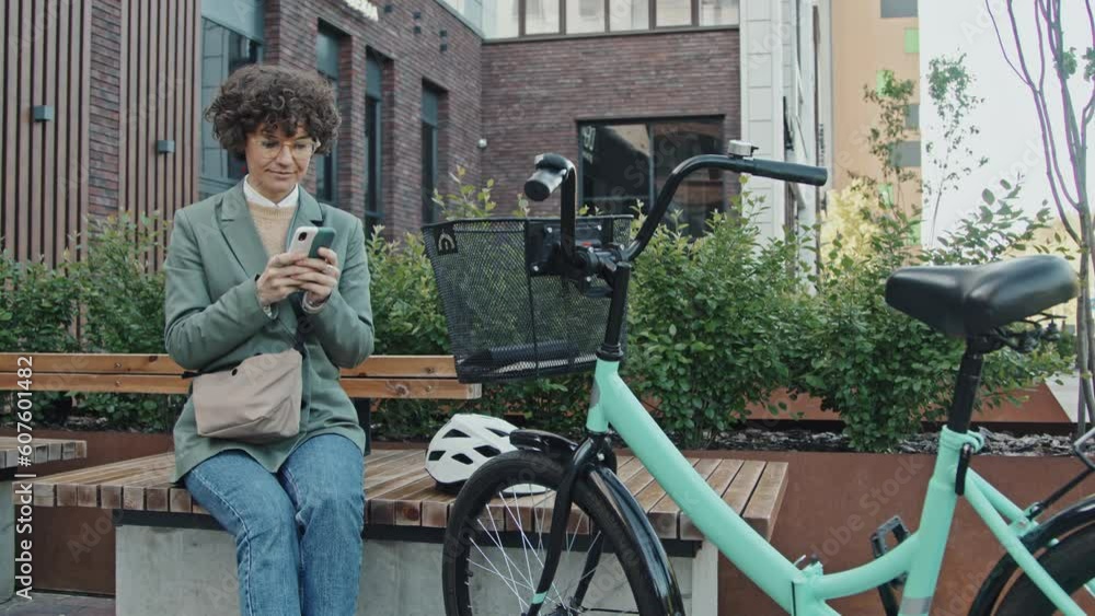 Medium long shot of modern Caucasian businesswoman sitting on bench with her bicycle nearby searching information in Internet on smartphone Medium long shot of modern Caucasian businesswoman sitting on bench with her bicycle nearby searching information in Internet on smartphone