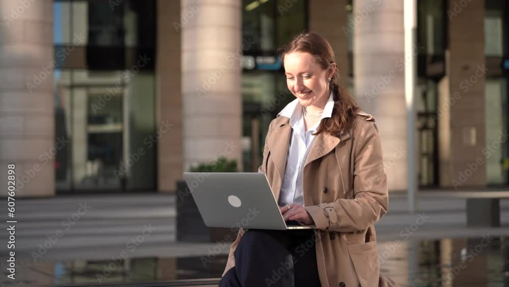 Smiling confident young woman freelancer in a trench coat working using a laptop typing text near the office. Smiling confident young woman freelancer in a trench coat working using a laptop typing text near the office.