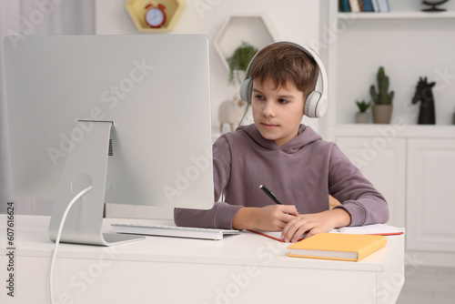 Boy writing in notepad while using computer and headphones at desk in room. Home workplace