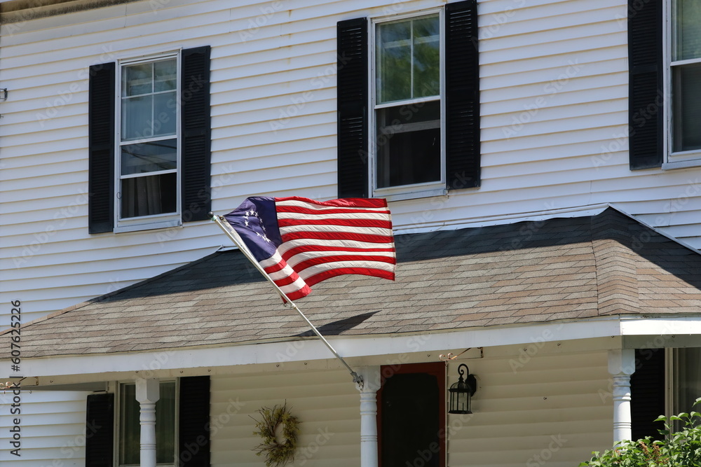 An American colonial flag with thirteeen stars representing the early colonies hanging from a wooden flagpoe in front of home with windows and shutters.