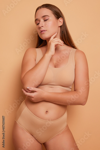 Portrait of beautiful young woman with long hair wearing bra and panties standing on background in the studio, with eyes down and hand up at her chin, body positivity concept, copy space