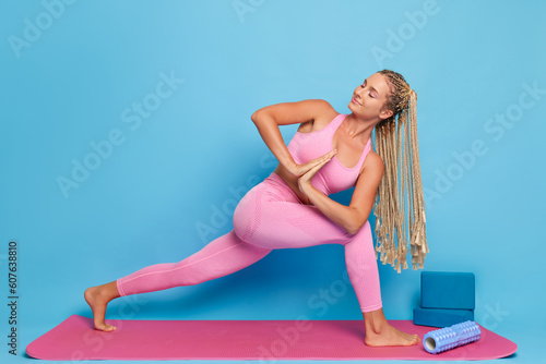 Sporty young woman in pink yoga costume doing workout on pink yoga mat standing in yoga pose against blue background, sport and fitness concept, copy space