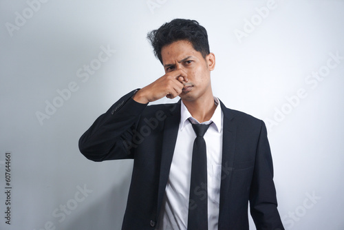 Portrait of bored or confused handsome Asian young man in suit standing, pinching his nose. indoor studio shoot, isolated on white background.