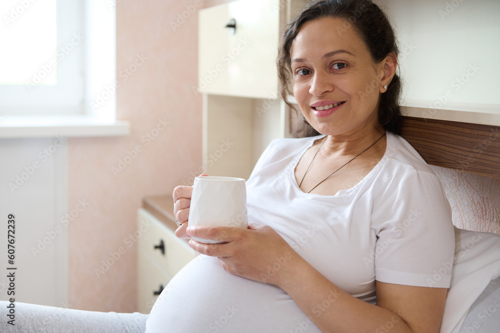 Beautiful multi ethnic adult pregnant woman in white t-shirt, cutely smiling looking at camera, relaxing on the bed, holding a big cup of healthy beverage. Happy moments of pregnancy. Expecting a baby Beautiful multi ethnic adult pregnant woman in white t-shirt, cutely smiling looking at camera, relaxing on the bed, holding a big cup of healthy beverage. Happy moments of pregnancy. Expecting a baby