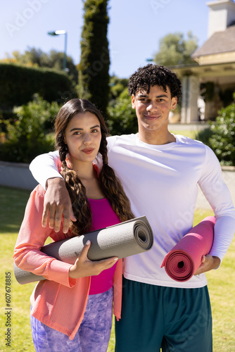 Portrait of confident biracial young couple with exercise mats standing in yard on sunny day