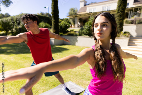 Confident biracial young couple practicing warrior 2 pose while standing in yard on sunny day