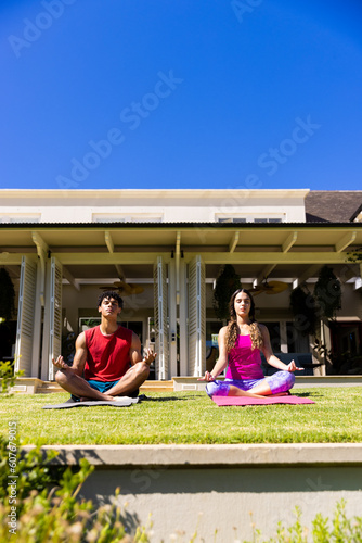 Biracial young couple practicing meditation while sitting in yard against house under clear blue sky