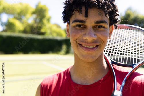 Close-up of biracial young man with tennis racket smiling in tennis court and looking at camera photo