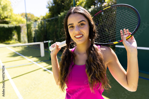 Portrait of smiling biracial beautiful woman holding tennis racket and standing in tennis court