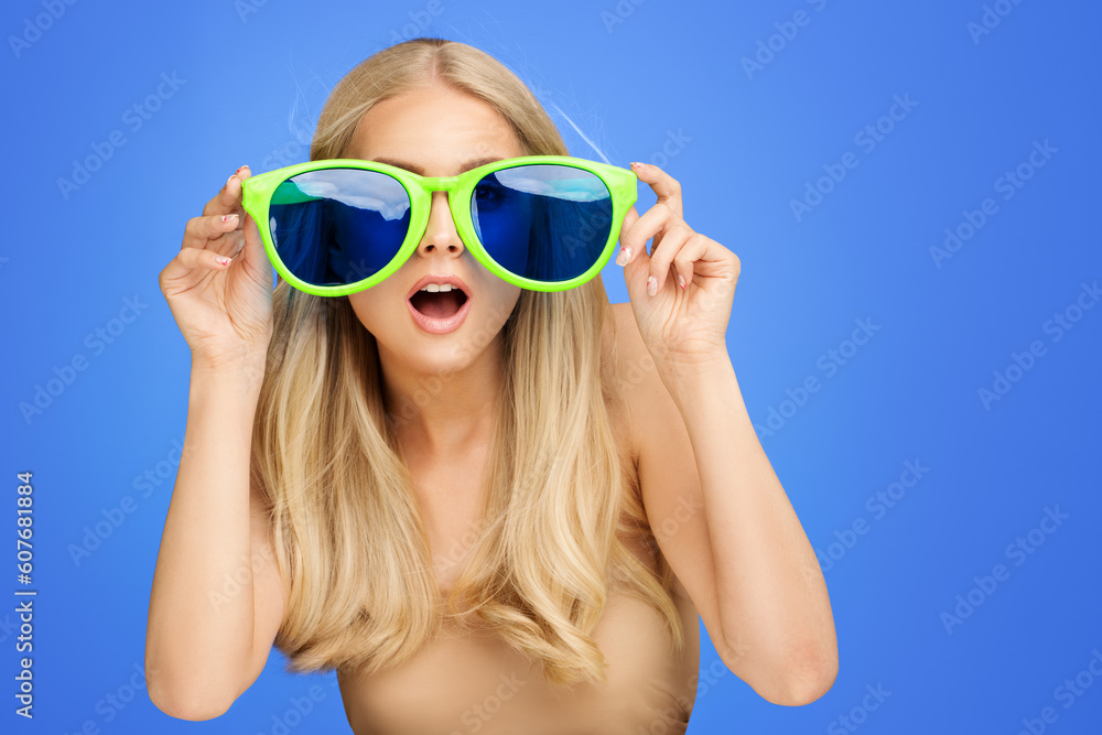 Surprised Girl in Big Party Glasses over Blue Background. Shocked Young Woman looking through Sunglasses. Portrait of Funny Girl Surprised Girl in Big Party Glasses over Blue Background. Shocked Young Woman looking through Sunglasses. Portrait of Funny Girl