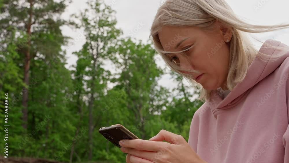 A young blonde woman uses a mobile phone while walking outdoors. A female blogger writes a message to her subscribers, talks about the benefits of walking and an active lifestyle. A young blonde woman uses a mobile phone while walking outdoors. A female blogger writes a message to her subscribers, talks about the benefits of walking and an active lifestyle.