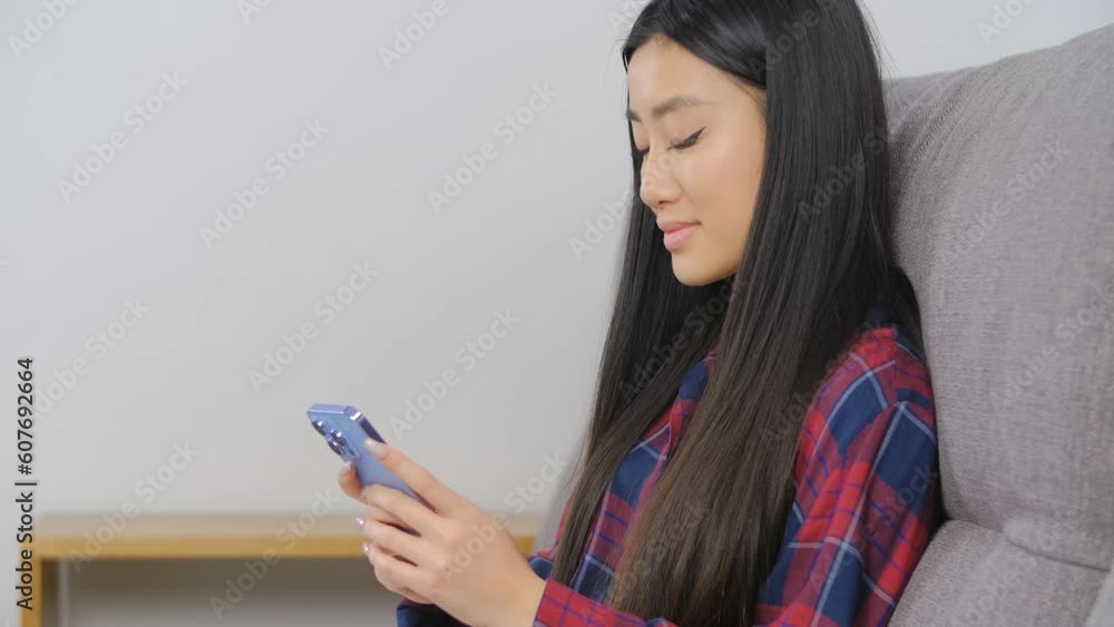 Cheerful Vietnamese female sitting on a couch in the living room and typing a message on a modern blue smart phone Cheerful Vietnamese female sitting on a couch in the living room and typing a message on a modern blue smart phone