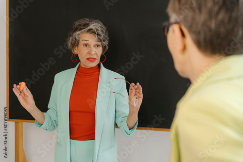 Woman in a bright suit standing in front of blackboard explaining something to another woman. photo