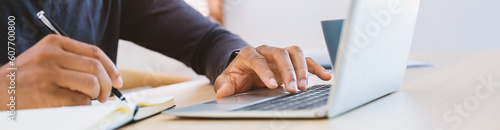 Closeup businessman hand holding pen writing on notebook, work on laptop computer in modern office. Student lecturing while studying online internet course. Write on journal diary banner (blur)
