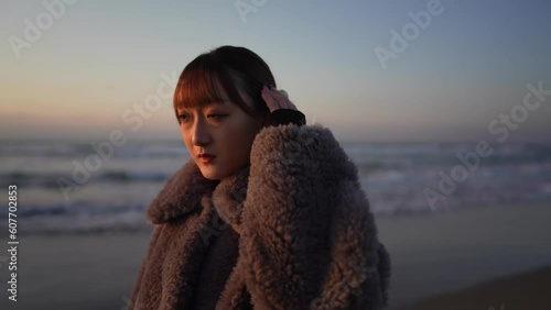 A young Japanese woman in her twenties, wearing a fluffy black coat, walking along the windy shoreline on a cold day at sunset on the sandy beach of Uchinada Town, Ishikawa Prefecture. photo