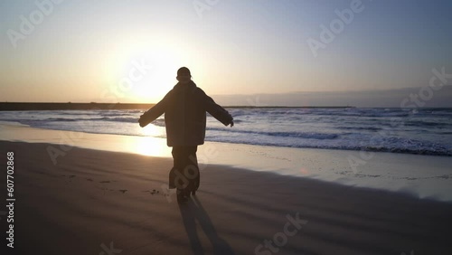 A young Japanese woman in her twenties, wearing a fluffy black coat, walking along the windy shoreline on a cold day at sunset on the sandy beach of Uchinada Town, Ishikawa Prefecture. photo