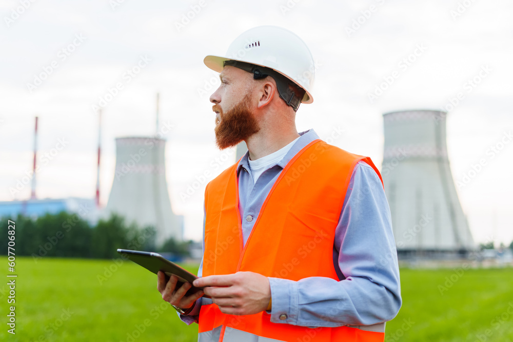 A professional engineer in a protective helmet and an orange safety vest holds a tablet in his hands. A man with a beard stands against the background of a power plant