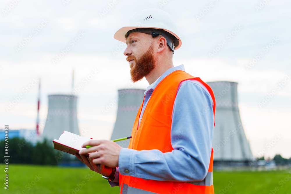 An engineer with a notebook on the background of a power plant. The man is wearing a white helmet and an orange vest An engineer with a notebook on the background of a power plant. The man is wearing a white helmet and an orange vest
