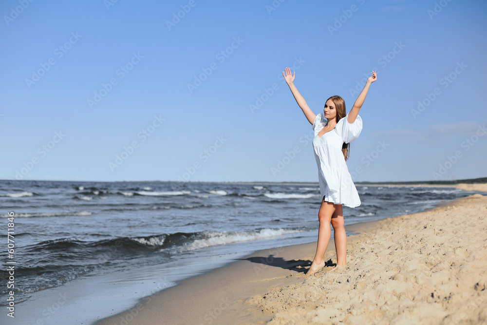 Happy, beautiful woman on the ocean beach standing in a white summer dress, raising hands.