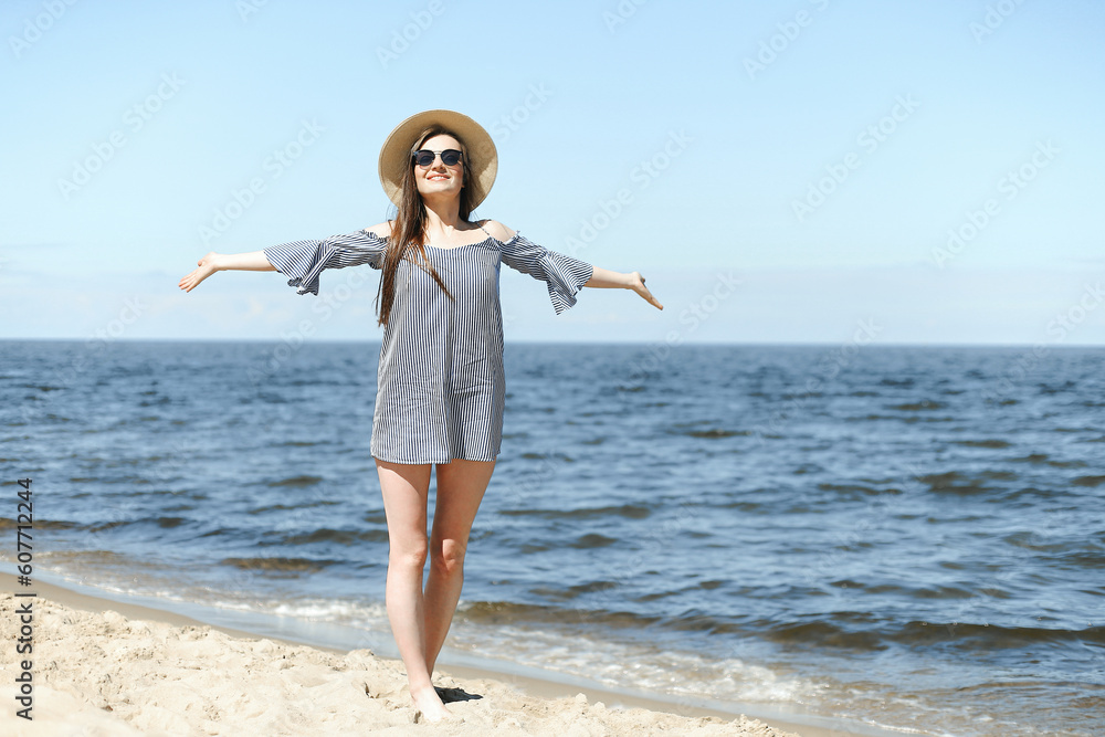Happy smiling woman in free bliss on ocean beach standing with open hands. Brunette female model in sunglasses and hat enjoying nature during travel holidays vacation outdoors, medium shot. Happy smiling woman in free bliss on ocean beach standing with open hands. Brunette female model in sunglasses and hat enjoying nature during travel holidays vacation outdoors, medium shot.