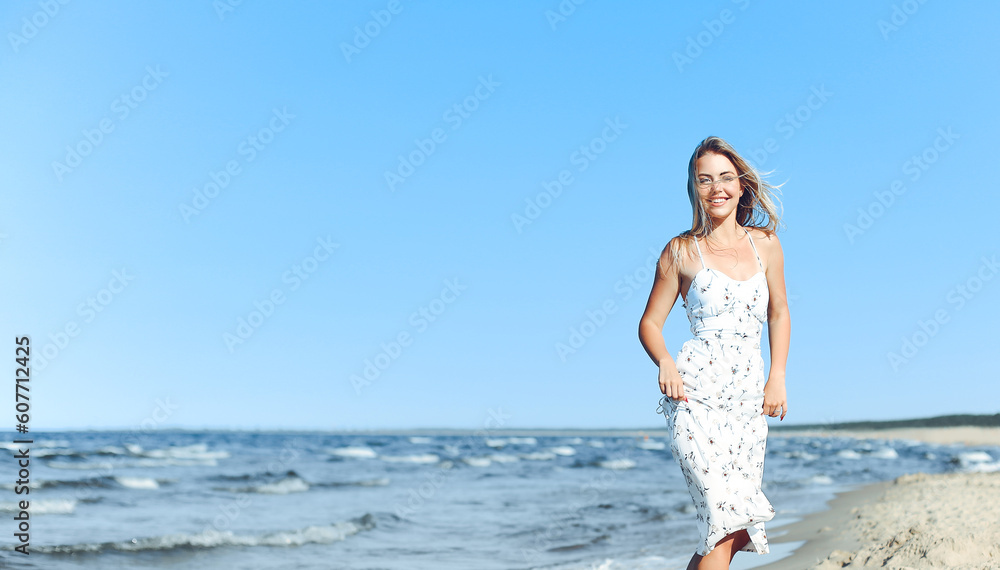 Happy blonde woman in free happiness bliss on ocean beach standing straight. Portrait of a female model in white summer dress enjoying nature during travel holidays vacation outdoors.