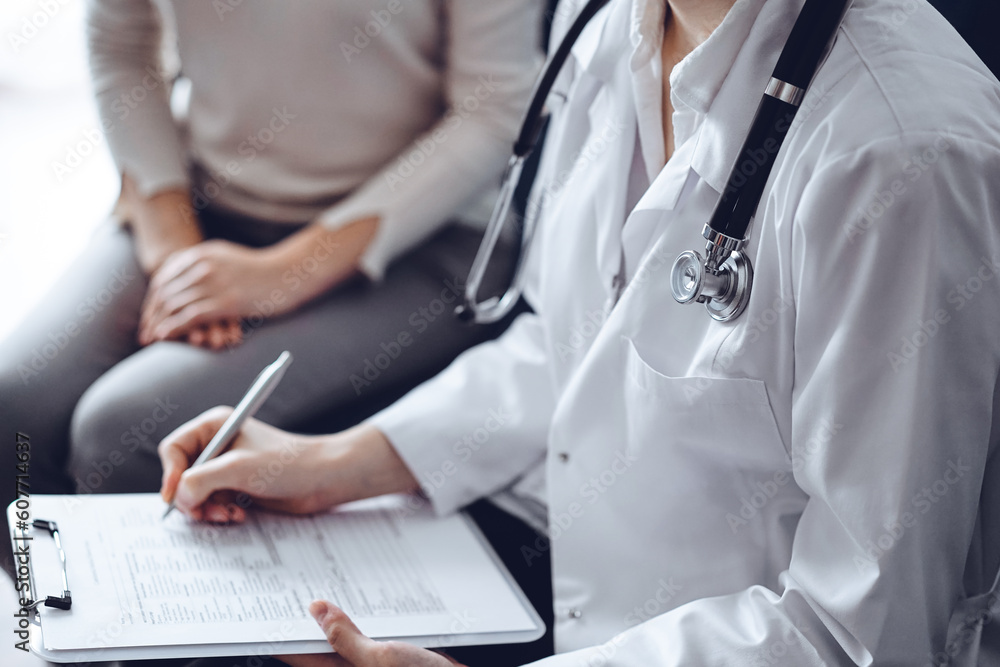Doctor and patient sitting at sofa in clinic office. The focus is on female physician's hands filling up the medication history record form, close up. Medicine concept. Doctor and patient sitting at sofa in clinic office. The focus is on female physician's hands filling up the medication history record form, close up. Medicine concept.