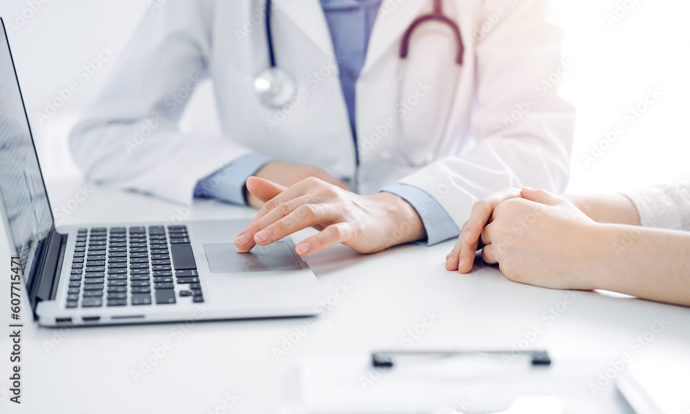 Doctor and patient sitting near each other at the desk in clinic. The focus is on female physician's hands using laptop computer, close up. Medicine concept. Doctor and patient sitting near each other at the desk in clinic. The focus is on female physician's hands using laptop computer, close up. Medicine concept.