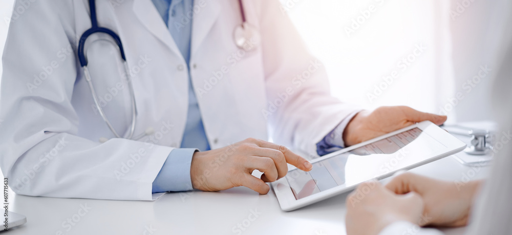 Doctor and patient sitting opposite each other at the desk in clinic. The focus is on female physician's hands pointing into tablet computer touchpad, close up. Medicine concept. Doctor and patient sitting opposite each other at the desk in clinic. The focus is on female physician's hands pointing into tablet computer touchpad, close up. Medicine concept.