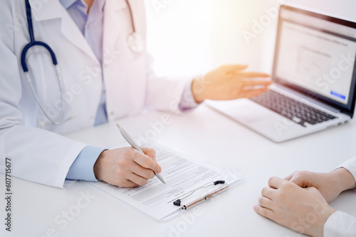 Doctor and patient sitting opposite each other at the desk in clinic. The focus is on female physician's hands filling up the medication history record form or checklist, close up. Medicine concept.