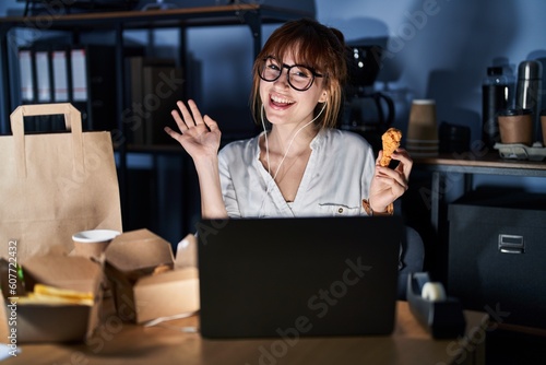 Young beautiful woman working using computer laptop and eating delivery food waiving saying hello happy and smiling  friendly welcome gesture