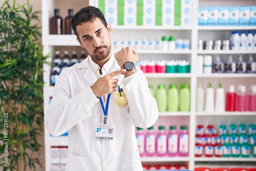 Handsome hispanic man working at pharmacy drugstore in hurry pointing to watch time, impatience, looking at the camera with relaxed expression © Krakenimages.com