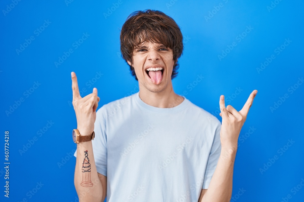 Hispanic young man standing over blue background shouting with crazy expression doing rock symbol with hands up. music star. heavy music concept. Hispanic young man standing over blue background shouting with crazy expression doing rock symbol with hands up. music star. heavy music concept.