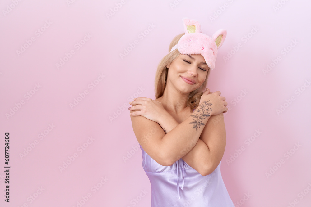 Young caucasian woman wearing sleep mask and pajama dress hugging oneself happy and positive, smiling confident. self love and self care
