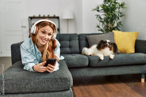 Young caucasian woman listening to music lying on sofa with dog at home