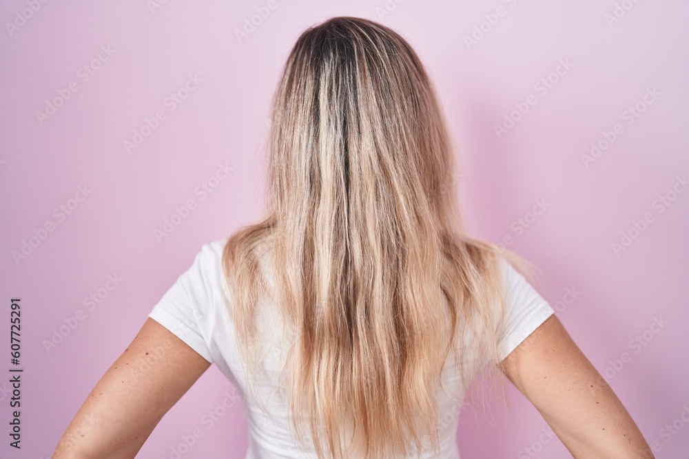 Young blonde woman standing over pink background standing backwards looking away with arms on body Young blonde woman standing over pink background standing backwards looking away with arms on body