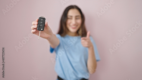 Young beautiful hispanic woman doing thumbs up pointing to key of new car over isolated pink background