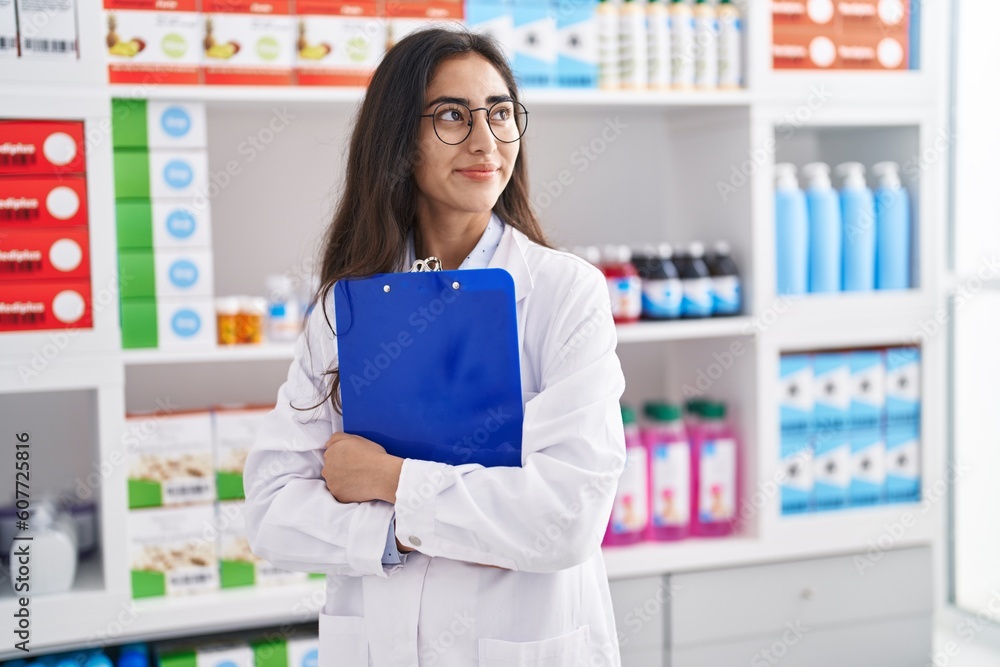 Young hispanic girl pharmacist hugging clipboard at pharmacy