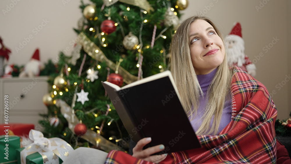 Young blonde woman reading book sitting on sofa by christmas tree at home