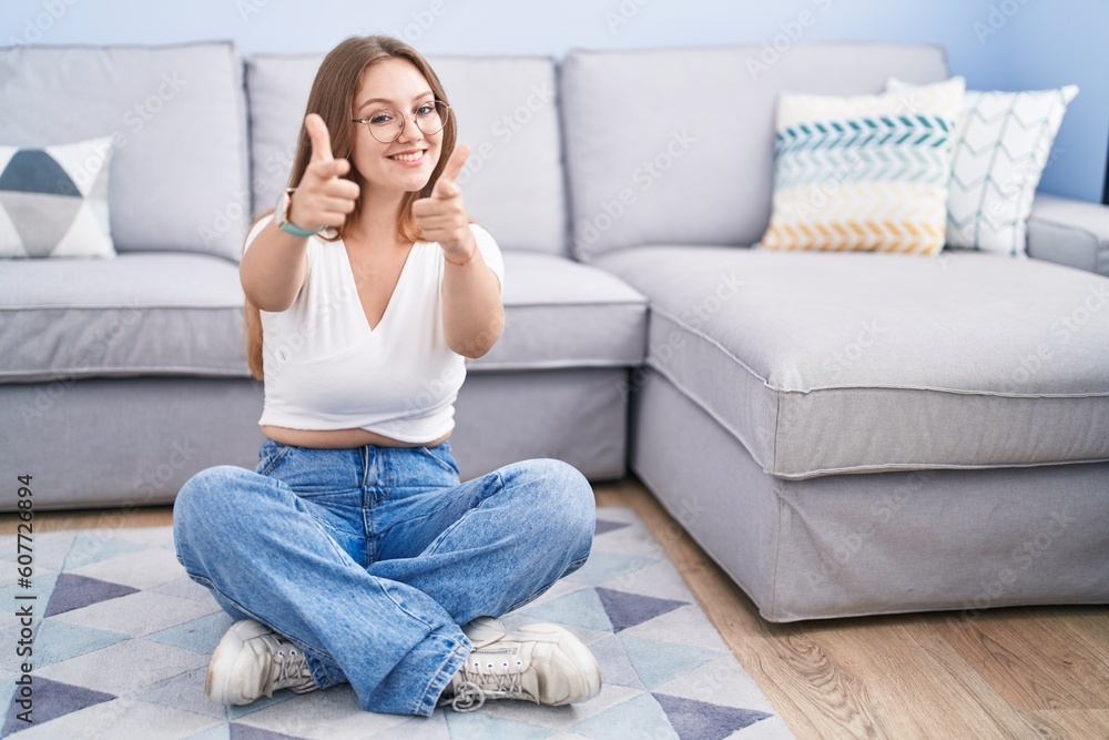 Young caucasian woman sitting on the floor at the living room pointing fingers to camera with happy and funny face. good energy and vibes. Young caucasian woman sitting on the floor at the living room pointing fingers to camera with happy and funny face. good energy and vibes.