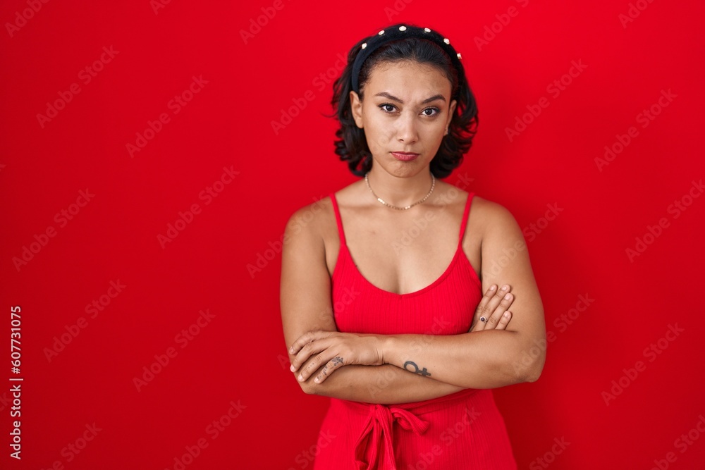 Young hispanic woman standing over red background skeptic and nervous, disapproving expression on face with crossed arms. negative person.