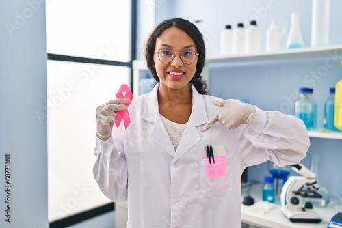 Young hispanic doctor woman working at scientist laboratory holding pink ribbon pointing finger to one self smiling happy and proud