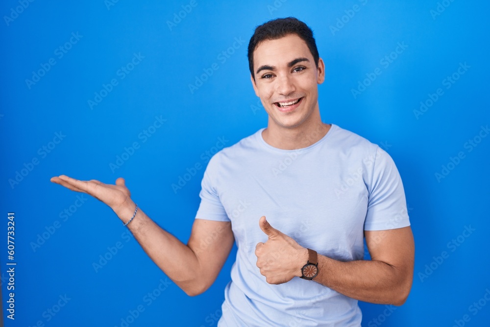 Young hispanic man standing over blue background showing palm hand and doing ok gesture with thumbs up, smiling happy and cheerful Young hispanic man standing over blue background showing palm hand and doing ok gesture with thumbs up, smiling happy and cheerful