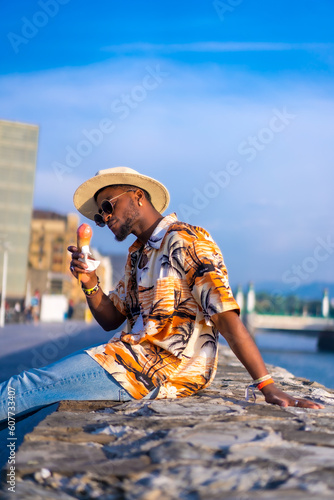 Portrait of a black ethnic man enjoy summer vacation at the beach eating an ice cream, sitting by the river in a vacation time